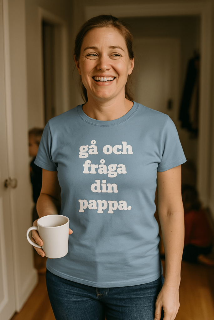 Kvinna i 30-årsåldern med blå t-shirt med texten “gå och fråga din pappa”, håller kaffekopp i hallen med barn i bakgrunden.